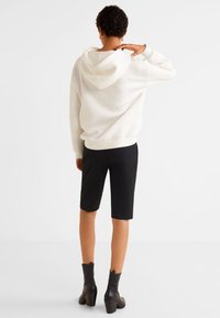 Sudadera blanca con capucha, pantalones cortos negros hasta las rodillas y botas de tacón negro a la altura del tobillo, vistas desde atrás, contra un fondo neutro.