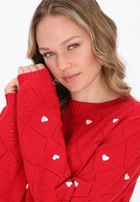 Pulover tricotat roșu cu un model în diamante și mici accente sub formă de inimioare albe. Mâneci lungi cu manșete în dungi și decolteu rotund.