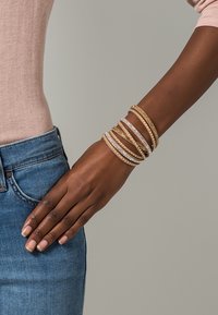 Guld- och silverarmband staplade på en handled, med släta och texturerade ytor, några med klara ädelstenar, mot en ljus bakgrund.