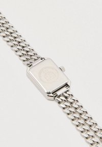 Srebrny metalowy zegarek na rękę z prostokątną kopertą, plecionym paskiem i teksturowanym tyłem z wyrytym logo oraz szczegółami.