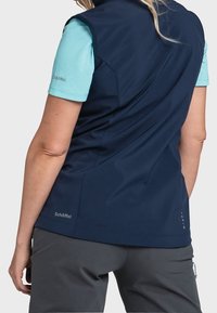 Námořnická bezrukávová bunda s světle modrou košilí pod ní. Odolná tkanina, hladká textura, jemné akcenty a logo značky viditelné na boku.