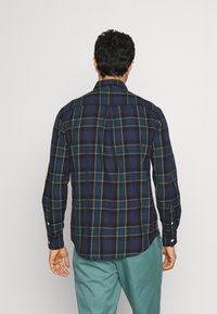 Hombre de espaldas, vistiendo una camisa de manga larga de cuadros en azul marino y verde, junto con pantalones en tono verde azulado, contra un fondo blanco liso.