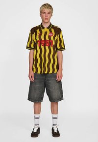 Brun- och gulrandig pikétröja med röd "1951"-tryck, kombinerad med mörka denimshorts. Vita strumpor med svarta ränder och mörka skor.