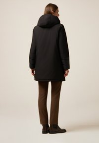 Abrigo negro acolchado con una capucha grande y estructurada, con mangas estrechas y detalles texturizados. Combinado con pantalones marrón de cuadros y botas.