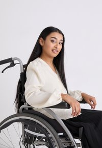 Blusa color crema con maniche a sbuffo, abbinata a una gonna nera plissettata. Modella seduta in una sedia a rotelle grigia con seduta texturizzata.