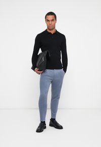 Schwarzes geripptes Langarmpolo, hellblaue schmal geschnittene Hose und schwarze Leder-Slipper; hält eine schwarze Lederjacke. Weißer Hintergrund.