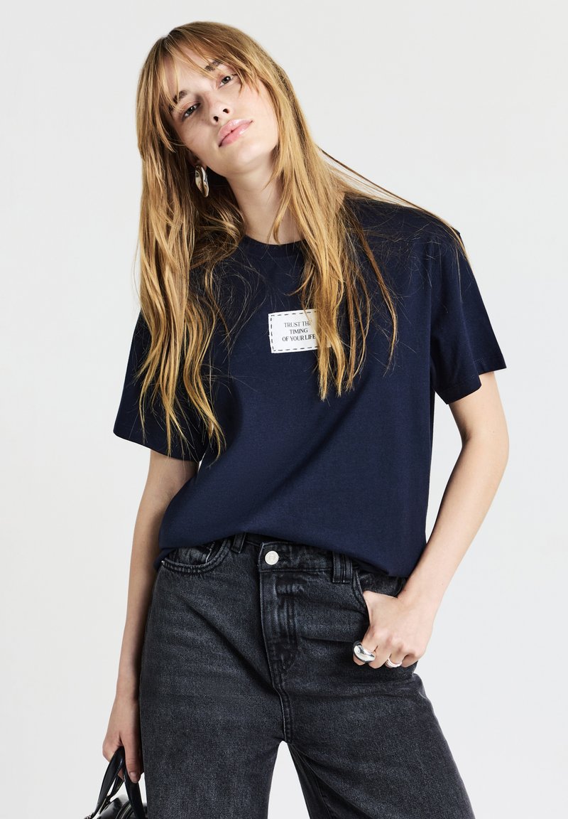 Giovane donna con lunghi capelli biondi, che indossa una maglietta blu scuro con un'applicazione di testo e jeans scuri, mentre tiene una borsa nera su uno sfondo bianco.