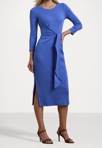 Blå jerseykjol med långa ärmar, en bunden midjedetalj och sid/slitsar. Åtföljd av bruna klackar med remmar mot en neutral bakgrund.