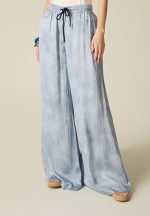 Frau trägt hellblaue, weit geschnittene Hose mit Kordelzug, kombiniert mit beigen Mokassins, steht vor einem einfarbigen Hintergrund.