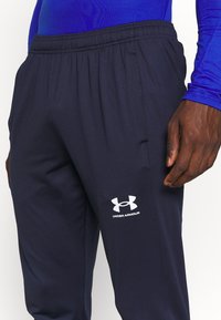 Navy urheiluhousut, jotka on valmistettu sileästä kankaasta ja joissa on elastinen vyötärö. Vasemmassa reidessä on valkoinen Under Armour -logo.