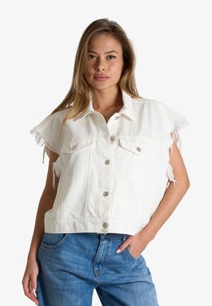 Giovane donna che indossa una giacca di jeans bianca senza maniche con bordi sfrangiati e jeans blu, posa con una mano in tasca.