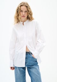 Weißes Hemd mit Stehkragen, das vertikale Faltendetails und lange, leicht puffige Ärmel aufweist. Kombiniert mit hellblauen Jeans.