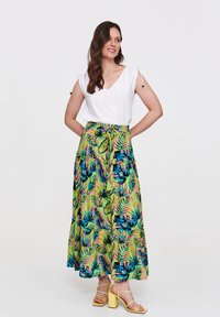 Biała bluzka z dekoltem w serek i krótkimi rękawami, zestawiona z długą, kolorową spódnicą z tropikalnym wzorem liści w odcieniach zieleni, niebieskiego i różowego. Żółte sandały na blokowym obcasie.