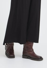 Rjave usnjene gležnjarje z zaobljenim nosom, ravnim podplatom in podrobnostjo z zaponko okoli gležnja, v kombinaciji z ohlapno črno obleko.