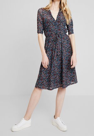 Kobieta ubrana w granatową, kwiecistą sukienkę do kolan z rękawami 3/4, dekoltem w serek i białe platformowe trampki, stojąca z jedną nogą wyprostowaną.