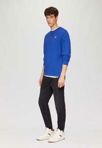Felpa blu con collo a giro, logo sobrio, abbinata a jeans neri e scarpe da ginnastica bianche con accenti rossi. Tessuto morbido e design aderente.