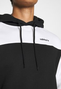 Svart och vit Adidas-hoodie med en delad design, som har en svart huva, snoddar och logotyp på bröstet. Mjuk tygtextur.