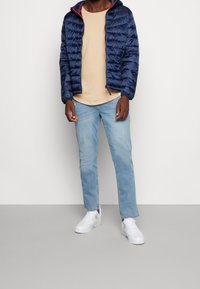 Giacca puffy blu navy con dettagli rossi, t-shirt beige e jeans azzurri chiari. Il modello indossa sneakers bianche, mostrando un outfit casual.