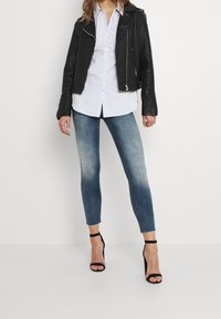Chaqueta de motorista de cuero negro, camisa blanca de botones y jeans ajustados de degradado azul claro con dobladillos deshilachados. Sandalias de tacón alto negras con tiras.