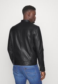 Zwarte leren jack met een gladde textuur, getailleerde pasvorm en staande kraag; voorzien van ritsmanchetten en naaddetails.