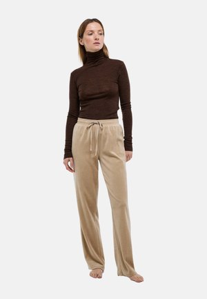 Femme debout pieds nus, portant un pull à col roulé marron et un pantalon de détente beige avec cordon de serrage, sur un fond uni.