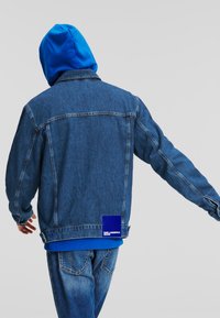 Jachetă din denim cu un hanorac albastru dedesubt. Jacheta are un patch pe spate cu branding și este într-o nuanță medie cu accente uzate.