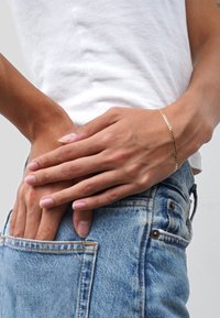 Gold chain bracelet resting on a hand, positioned near the waist. Worn with light blue denim jeans and a plain white t-shirt.