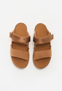 Brune sandaler laget av semsket skinn og stoff, med to brede stropper og en teksturert såle. Designet er åpent foran og flatt.