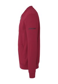 Sudadera roja de tela suave con cierre de cremallera, bolsillos laterales y un logo negro en la manga izquierda. Diseño simple y atlético.