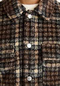 Zbliżenie brązowej i beżowej kratkowanej wełnianej kurtki z srebrnymi zatrzaskami na kremowym swetrze plecionym.