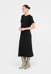 Rochie neagră, până la genunchi, cu mâneci scurte, având accente din nasturi pe o umăr și o textură netedă. Asortată cu cizme metalice cu imprimeu de șarpe.