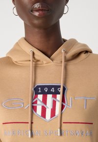 Bež hoodie s vezicama, izvezenim logotipom s crveno-bijelo-plavim štitom i tekstom "American Sportswear" na rubu.