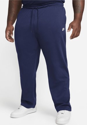 Uomo con pantaloni della tuta Nike blu navy e sneakers bianche, in piedi con le braccia rilassate lungo i fianchi su sfondo bianco.