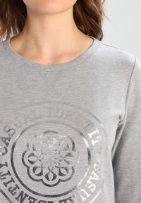 Felpa grigia chiara con una grafica rotonda in rilievo color argento. Presenta un colletto a girocollo a coste e maniche testurizzate.