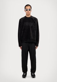 Μαύρο, απαλό φούτερ με την ένδειξη "VERSACE JEANS COUTURE" σε τονικές γραμματοσειρές, συνδυ�ασμένο με μαύρες φόρμες και γυαλιά ηλίου με άσπρο πλαίσιο, όρθια φιγούρα.