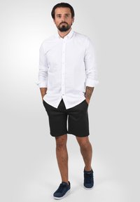 Weißes Hemd mit hochgekrempelten Ärmeln, schwarze Shorts und marineblau Sneakers; Tattoos sichtbar am Unterarm; neutraler Hintergrund.