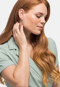 Boucles d'oreilles en argent portées par une personne aux longs cheveux ondulés châtaigne. Ils touchent leur cou, habillés d'un haut vert doux.