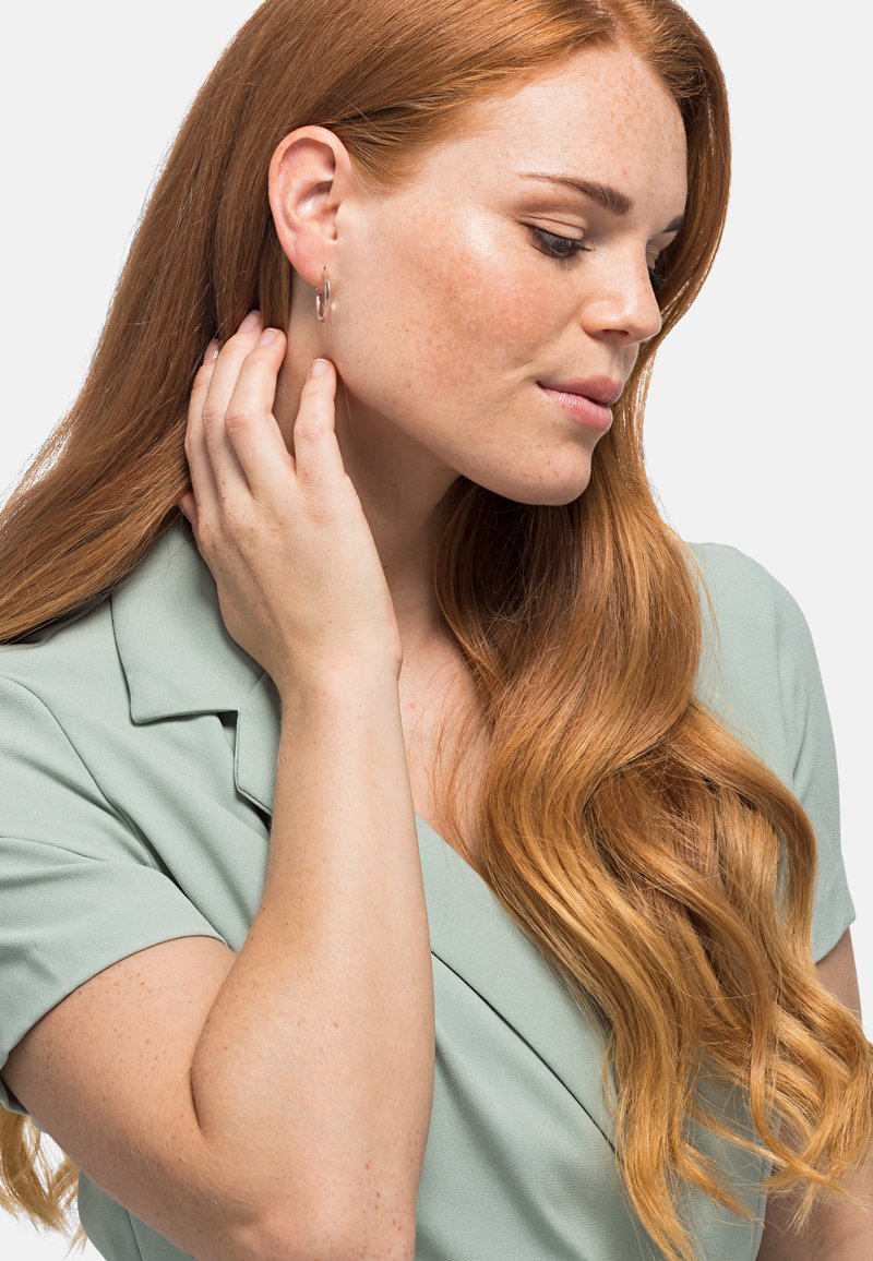 Boucles d'oreilles en argent portées par une personne aux longs cheveux ondulés châtaigne. Ils touchent leur cou, habillés d'un haut vert doux.