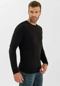 Uomo con capelli corti castani e barba, che indossa un maglione nero a maniche lunghe e jeans blu, che guarda a sinistra su uno sfondo bianco.
