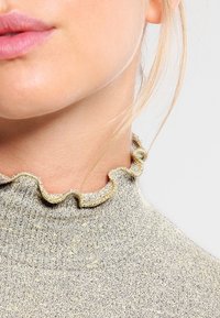 Helles, beiges Strickoberteil mit strukturierter Oberfläche, das über einen Rüschensaum und dezente metallische Fäden verfügt, die durch das Material gewoben sind.