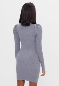 Kvinna med rakt, mörkt hår som bär en figurnära, långärmad, ribbad grå stickad klänning med puffade axlar, stående mot en otrevlig bakgrund.