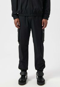 Sorte cargo-bukser med elastisk talje og mansjetter, der har to sidelommer og en glat, let stofstruktur. Båret med sorte sneakers.