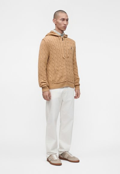 Άνδρας στέκεται φορώντας μια καφέ ζακέτα με φερμουάρ πλεκτή σε σχέδιο καλωδίου, ανοιχτόχρωμο ριγέ πουκάμισο, λευκό παντελόνι και μπεζ αθλητικά παπούτσια, σε απλό λευκό φόντο.