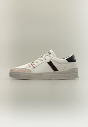 Baskets basses blanches avec embout beige, languette de talon noire, bande latérale noire avec surpiqûres rouges et lacets blancs sur fond gris.