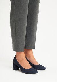 Granatowe zamszowe szpilki z okrągłym czubkiem i blokowym obcasem, noszone z dopasowanymi szarymi spodniami, prezentujące czystą i elegancką sylwetkę.