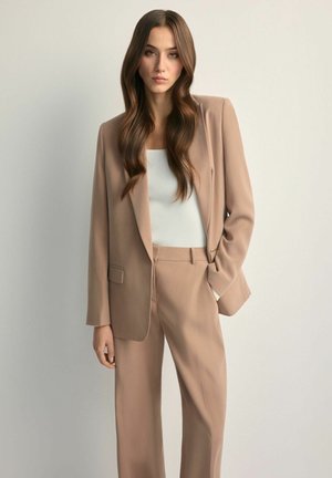 Młoda kobieta o długich, falowanych, brązowych włosach, ubrana w beżowy garnitur i białą bluzkę, stojąca na tle jednolitego, jasnoszarego tła.