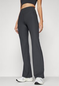 Pantalons ribbed taille haute de couleur gris foncé avec une coupe évasée, fabriqués à partir d'un tissu extensible, présentés avec un modèle portant des baskets blanches.