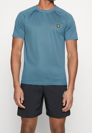 Uomo che indossa una maglietta sportiva a maniche corte blu con logo di uccello giallo e pantaloncini neri Adidas, in piedi contro un semplice sfondo bianco.