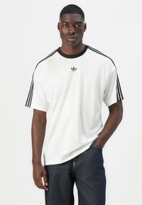 Witte T-shirt met zwarte schouderstrepen, met het Adidas-logo op de borst. De stof lijkt glad en heeft een relaxte pasvorm.