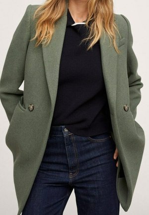 Femme portant un manteau vert cintré ouvert par-dessus un pull en tricot noir et un jean bleu foncé, les mains dans les poches du manteau.